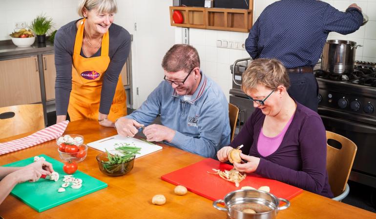 Bewoners snijden groente aan tafel terwijl de kookdame toekijkt en praat