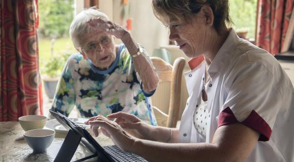 Thuiszorgmedewerker werkt op een tablet bij een oudere cliënt thuis en maakt zo gebruik van e-health.