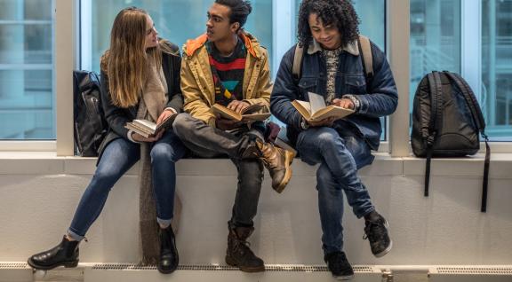 Drie jongeren op een vensterbank lezen een boek en praten met elkaar