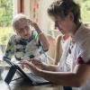 Thuiszorgmedewerker werkt op een tablet bij een oudere cliënt thuis en maakt zo gebruik van e-health.